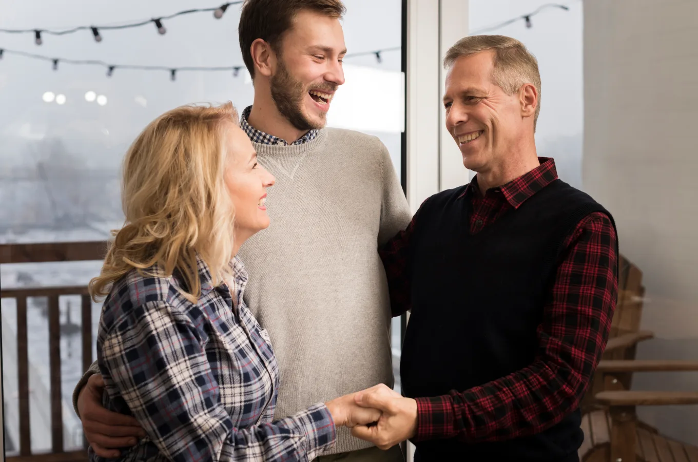Three adults standing together indoors, an older couple and a younger man, laughing, with string lights behind them