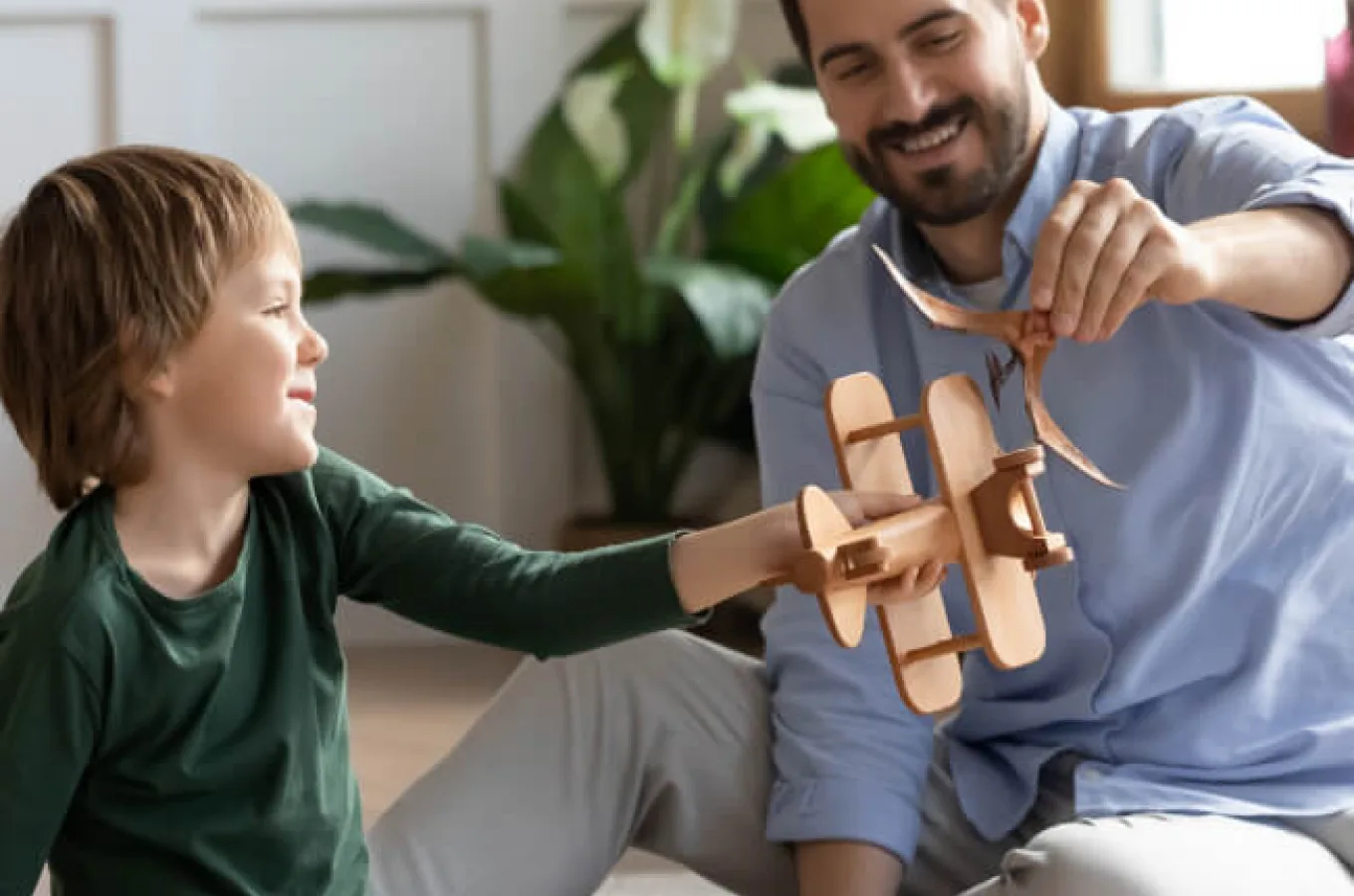 A smiling man and a young boy sitting on the floor, holding a wooden toy airplane between them, with a houseplant behind