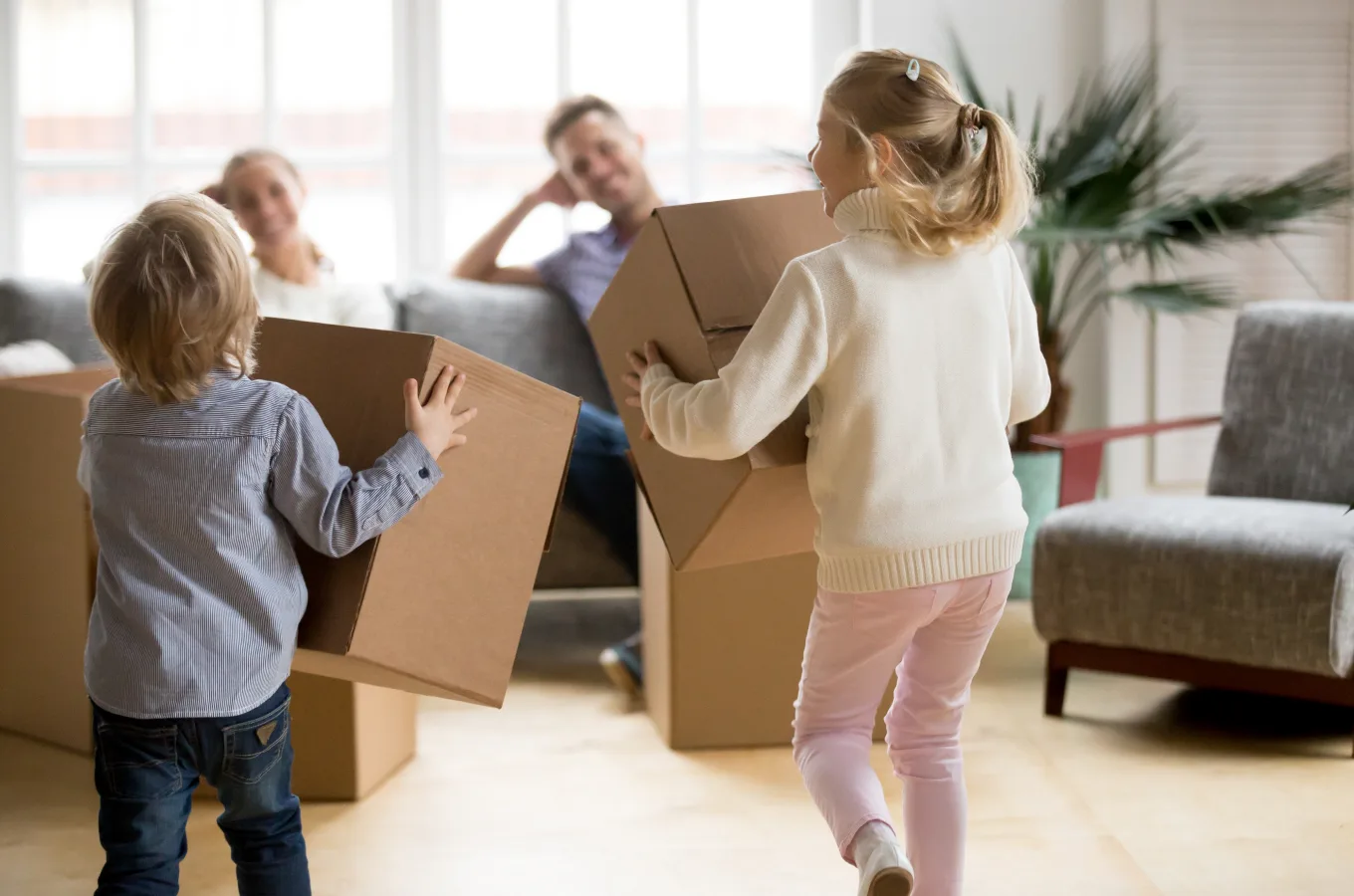 Two young children carrying cardboard moving boxes across a living room while two adults sit behind them