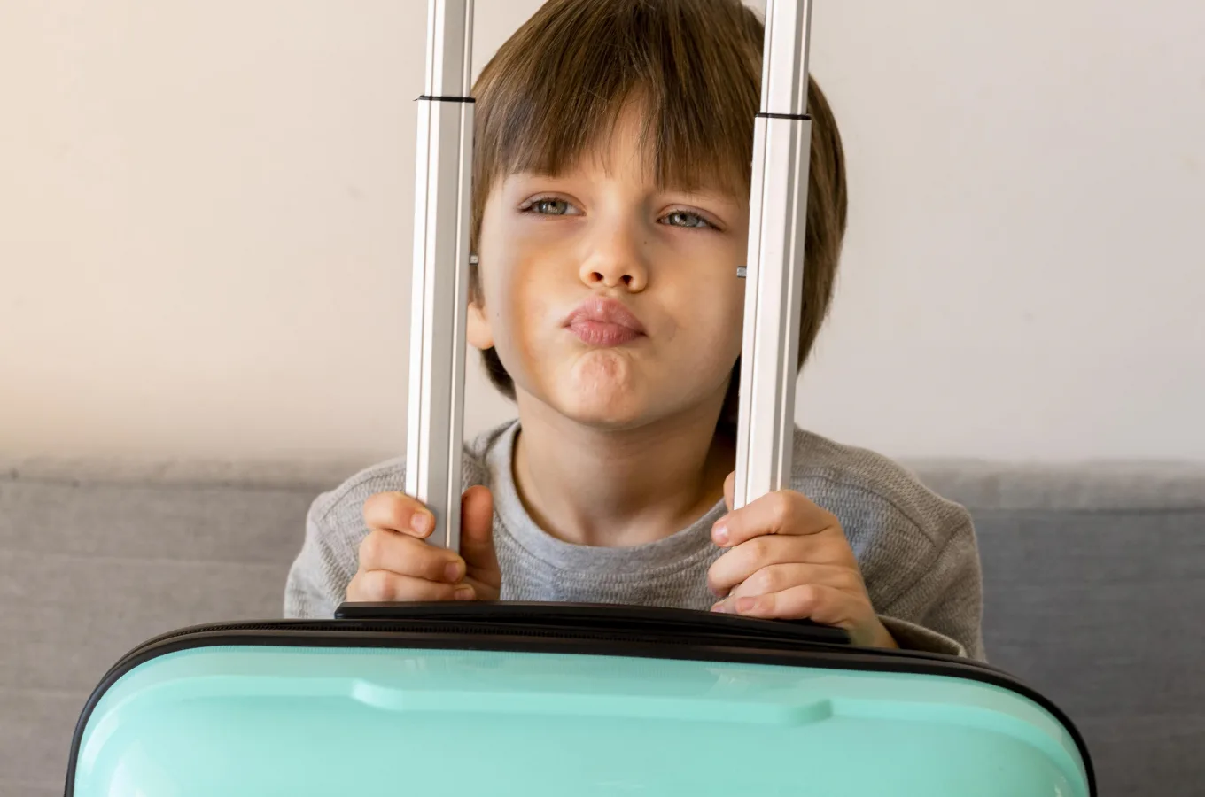 A child standing behind a teal suitcase, holding its extended handle with both hands and puffing out their cheeks