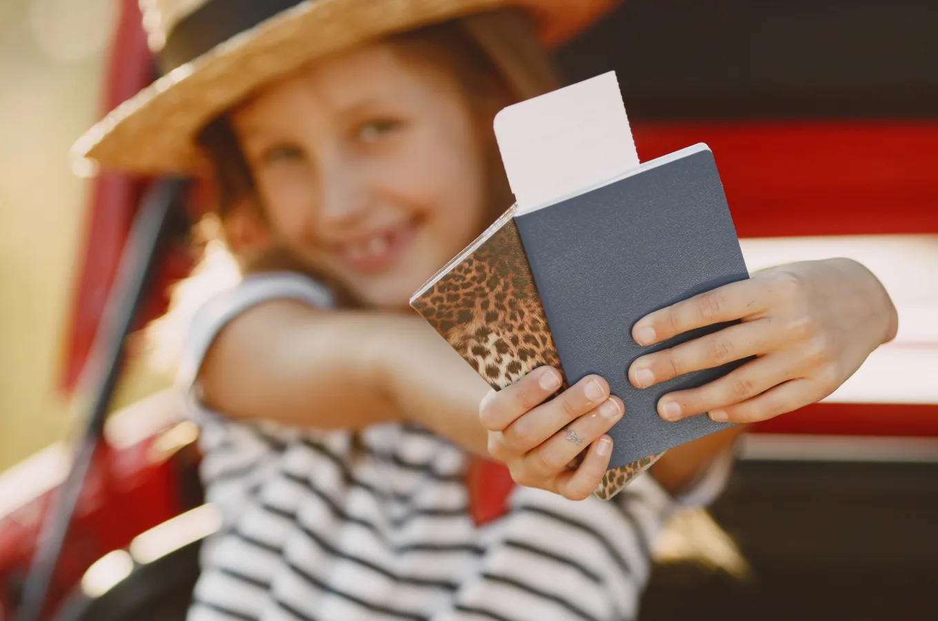 A girl in a straw hat holding out a passport with a boarding pass tucked inside, a red car behind her