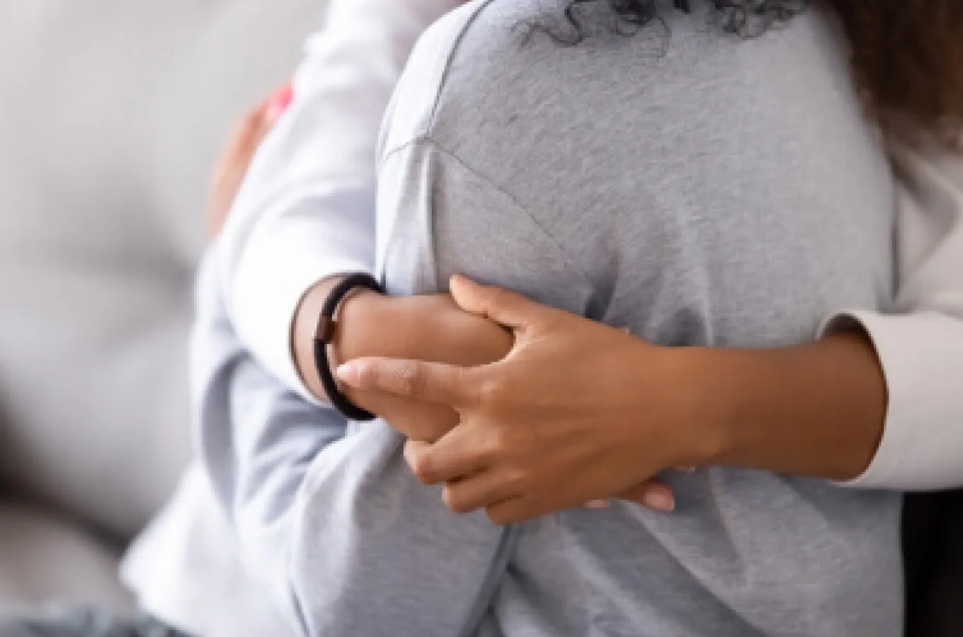 A child's arms wrapped around an adult from behind, hands clasped over a grey sweatshirt