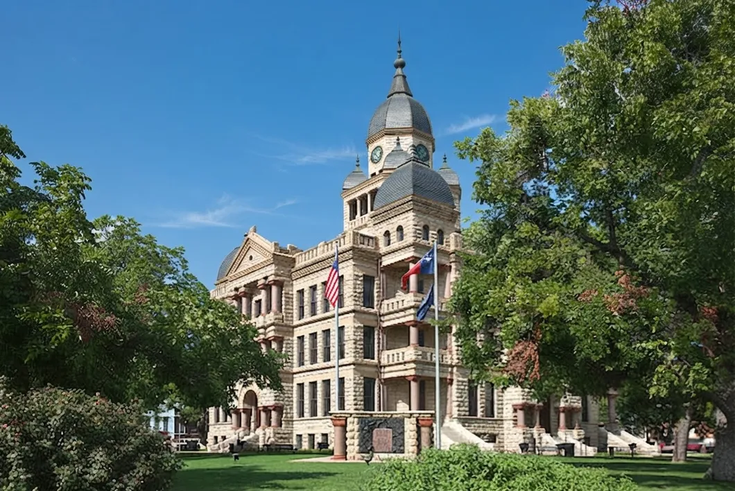 The historic Denton County courthouse, a pale stone building with a tall clock tower and flags, framed by leafy trees.