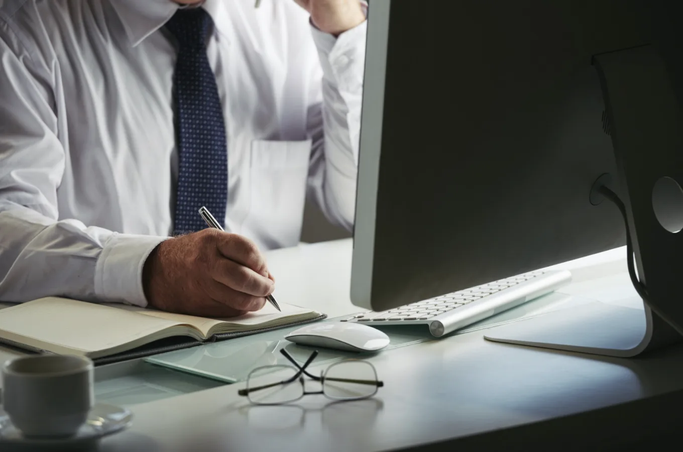 A person in a shirt and tie writing in a notebook at a desk, beside a desktop monitor, keyboard, glasses and a mug.