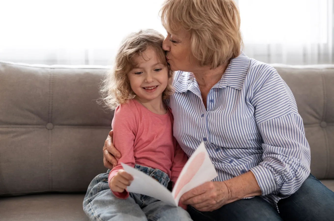 An older woman kissing a young girl on the forehead as they sit together on a sofa with an open picture book.