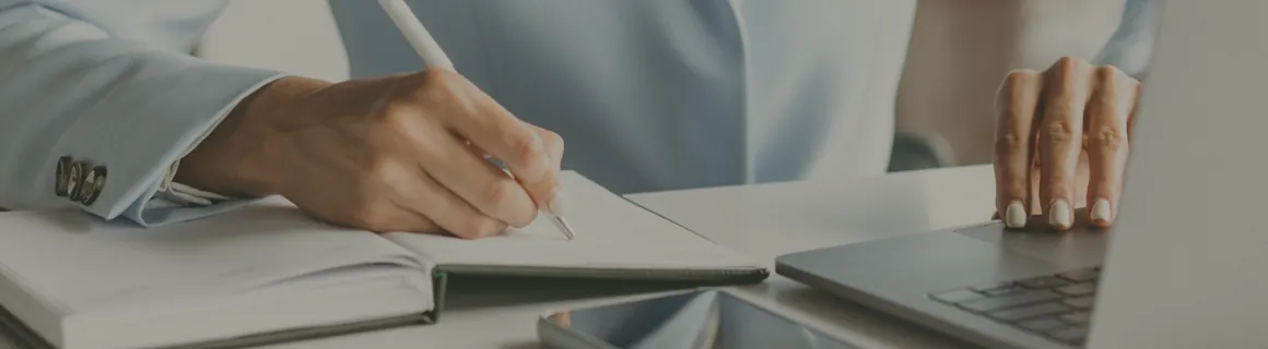 Hands writing in a notebook beside an open laptop