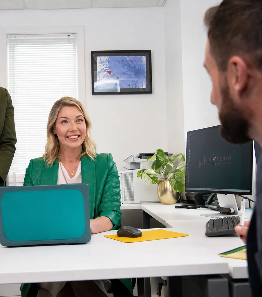 A smiling woman at a desk with a laptop, talking with a man in the foreground