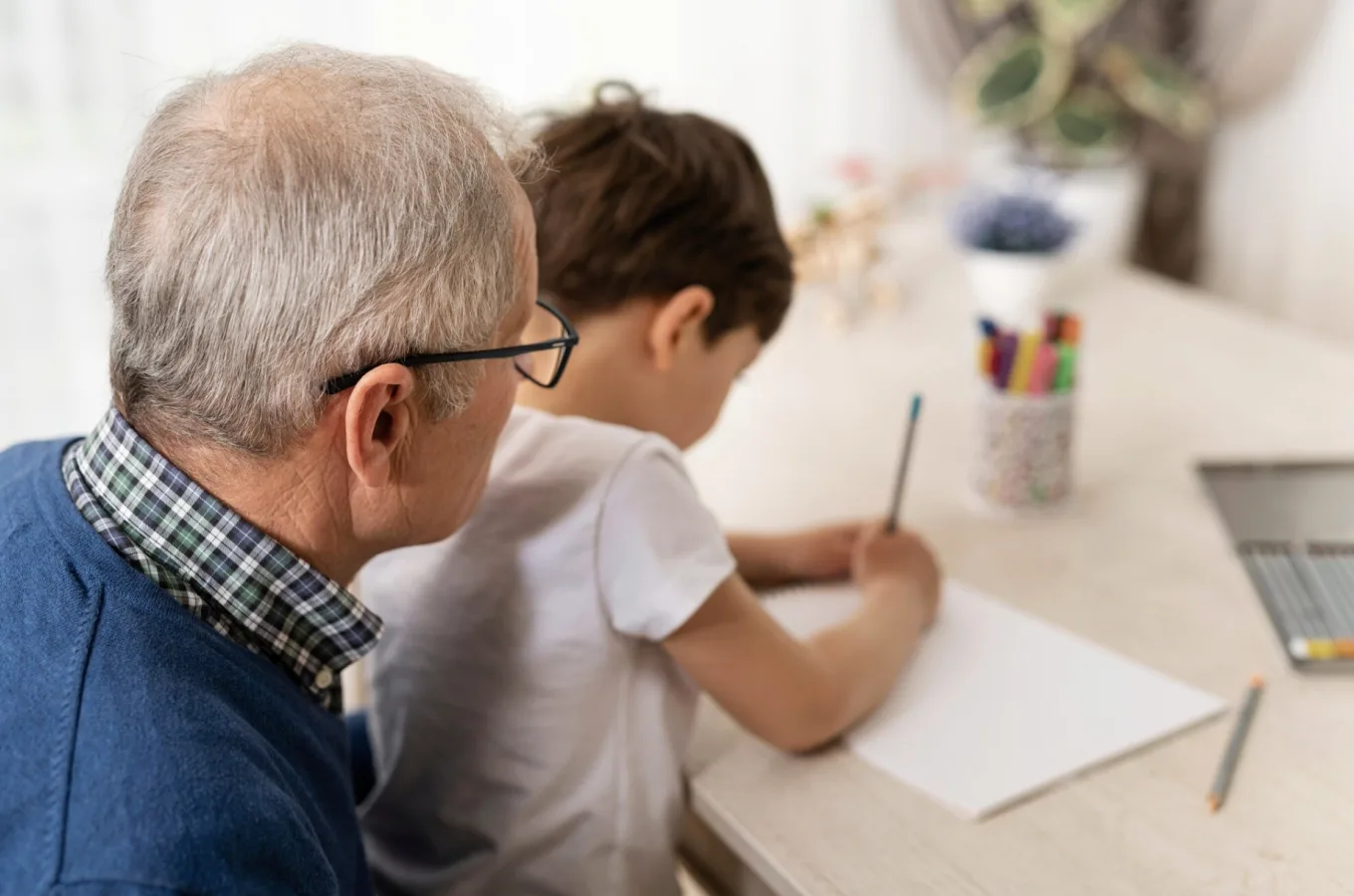 An older man in glasses leaning in beside a young boy who is drawing at a table, with colored pencils and a laptop nearby