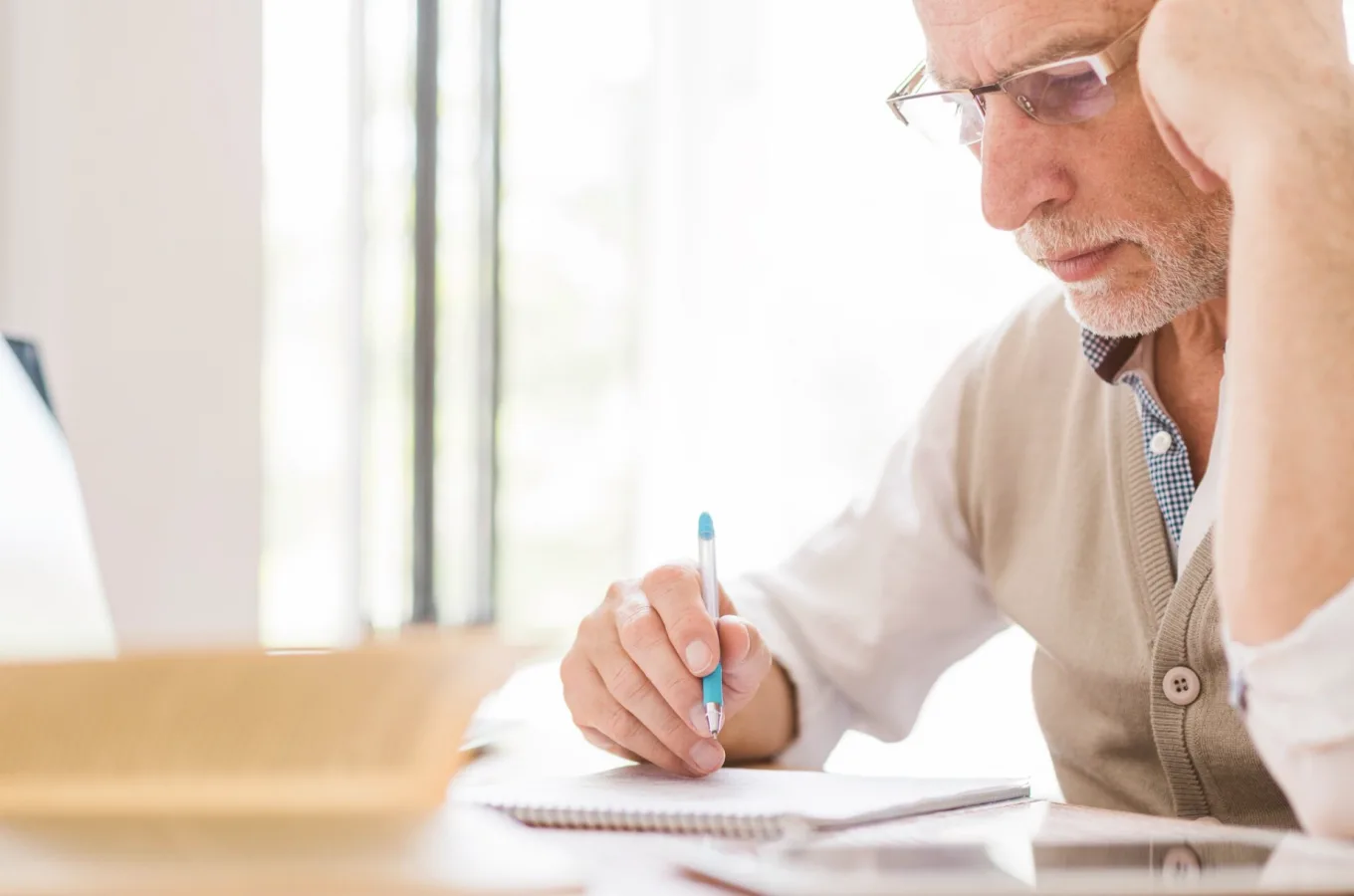 An older man in glasses resting his head on one hand while writing in a spiral notebook at a desk beside a bright window
