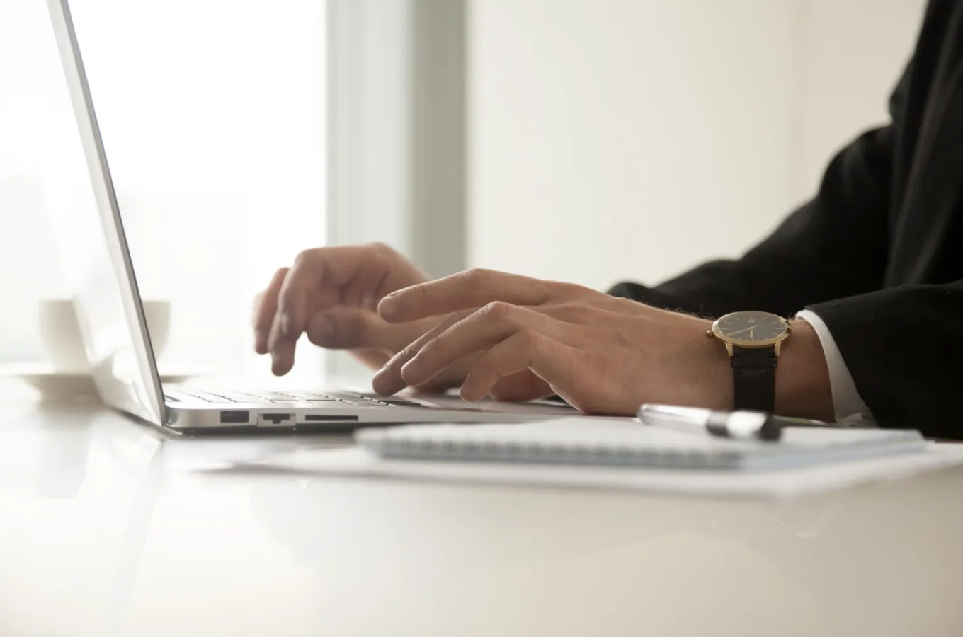 A person's hands typing on a laptop at a white table, with a notepad, a pen and a wristwatch in view.