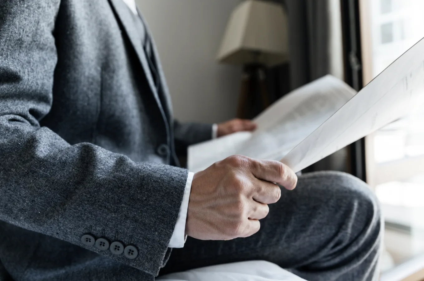 A person in a grey suit seated by a window, reading a printed document.