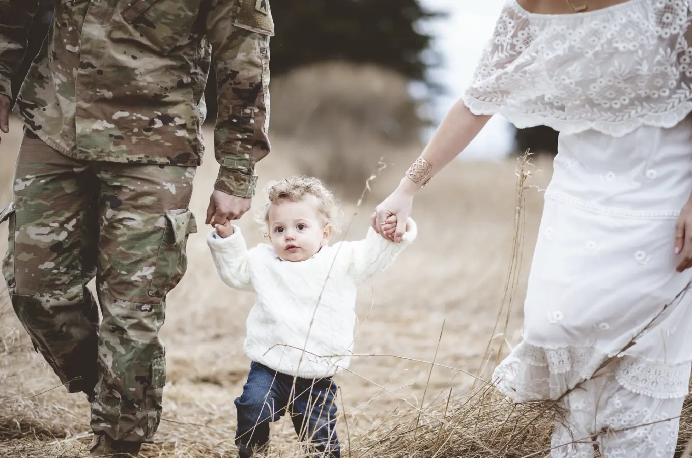 A person in camouflage uniform and a person in a white dress walk through dry grass holding a small child by the hands