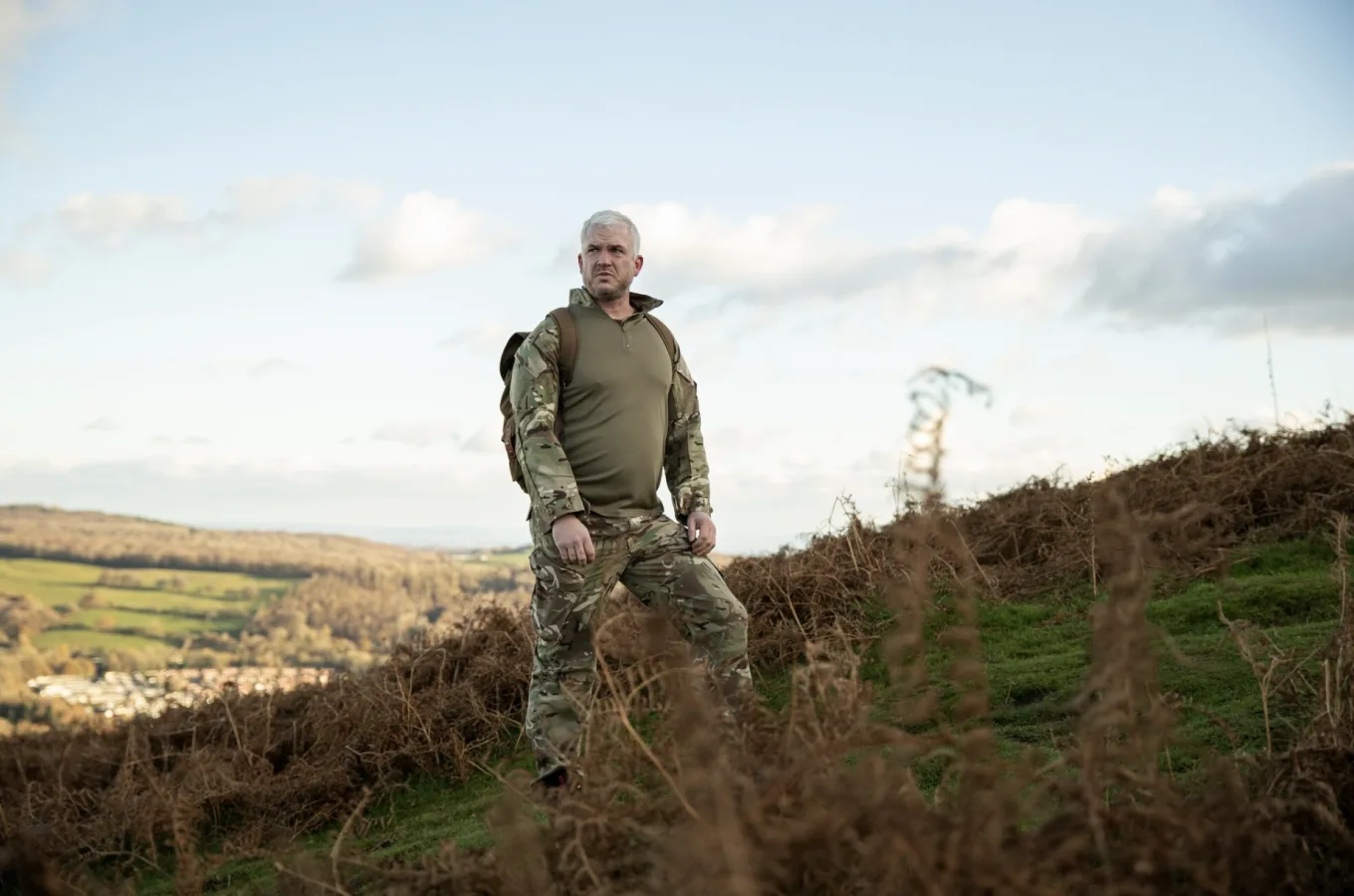 A person in camouflage uniform with a pack standing on a grassy hillside above a wooded valley