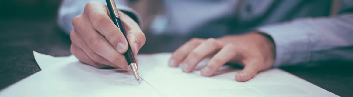 A hand signing a paper document with a pen at a desk