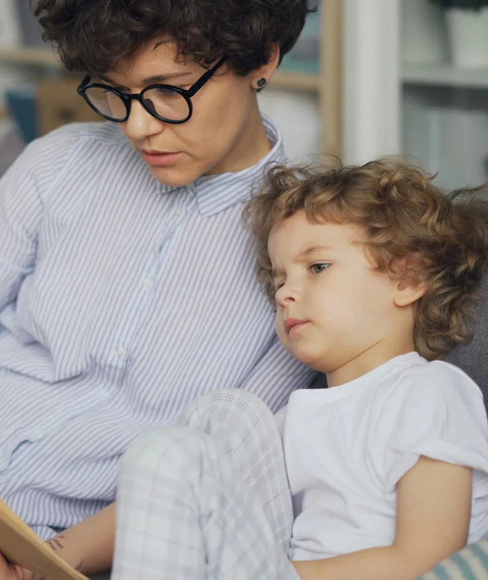 An adult in glasses and a small child sitting close together, looking down at a book