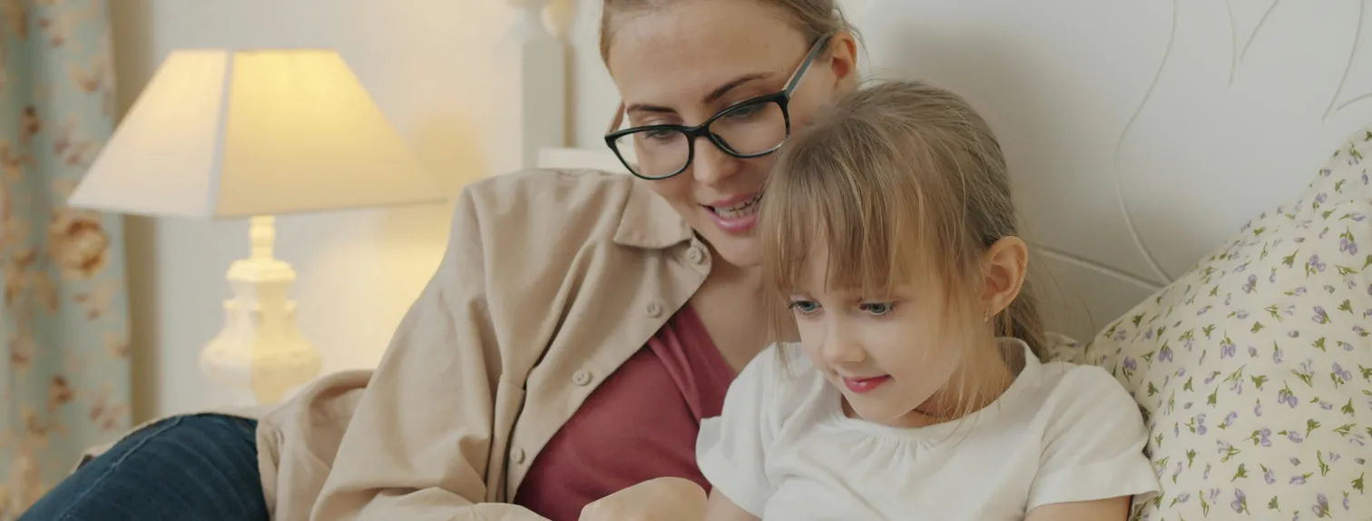 An adult in glasses and a child reading together beside a bedside lamp