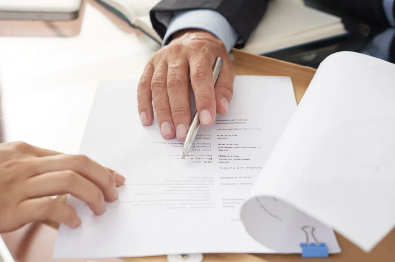 Two people's hands over printed documents on a desk, one of them holding a pen above a page