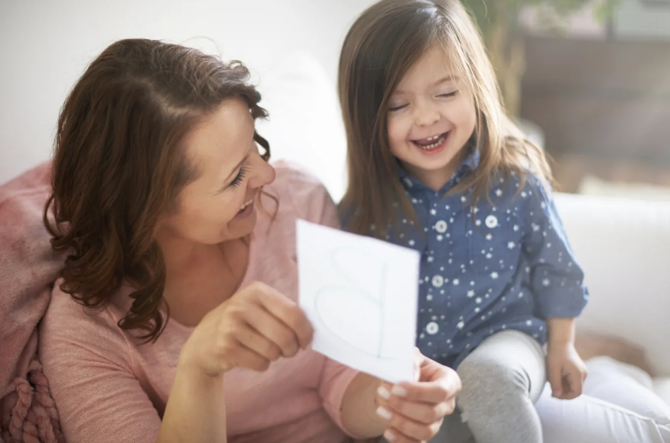 A woman and a laughing young girl sit together on a sofa holding up a sheet of paper with a drawing on it