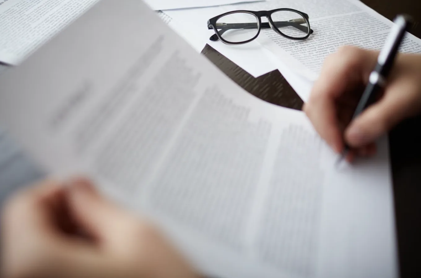 A hand holding a pen over printed pages, with a pair of eyeglasses resting on the papers