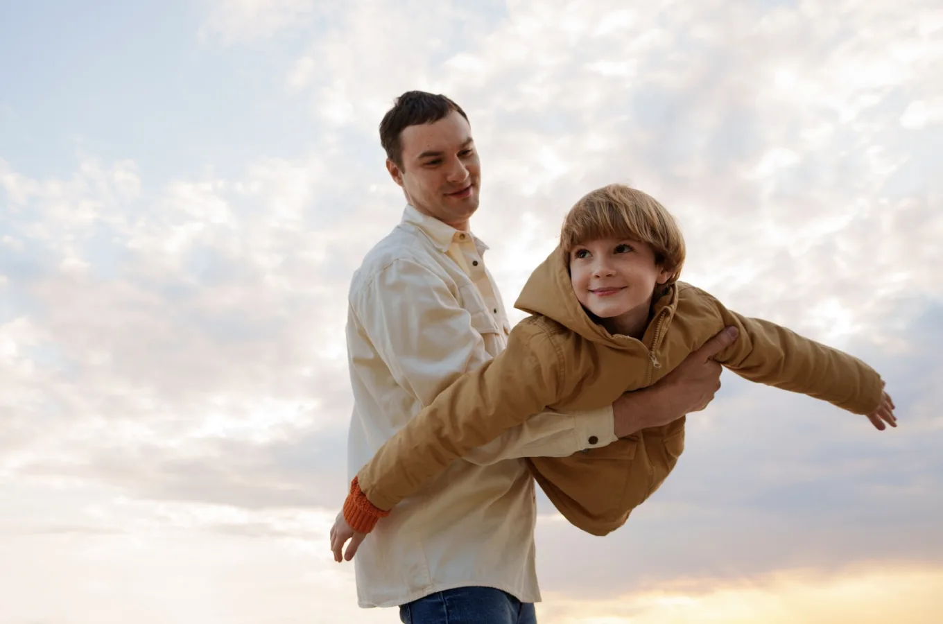 A man holding a young child aloft against a cloudy sky, the child's arms spread wide