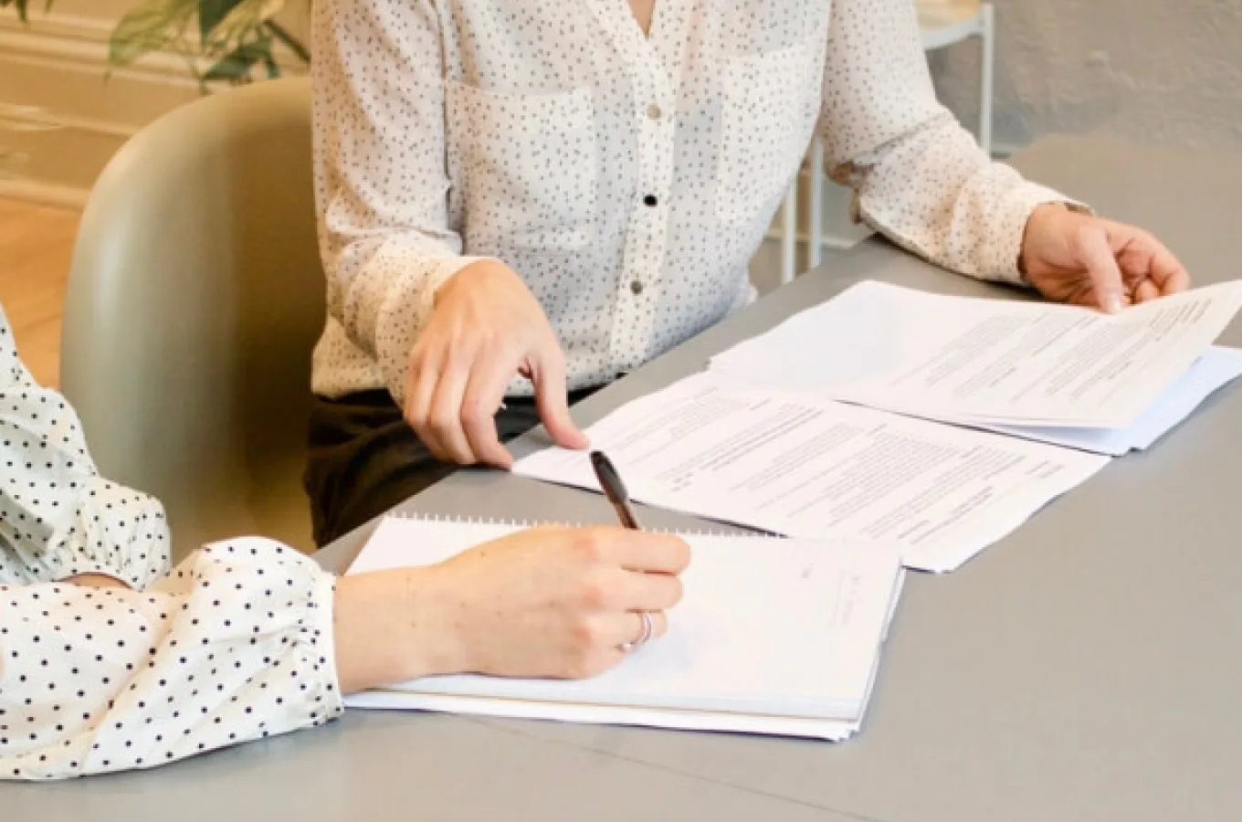 A woman at a table pointing toward printed documents while another person writes in a spiral notebook