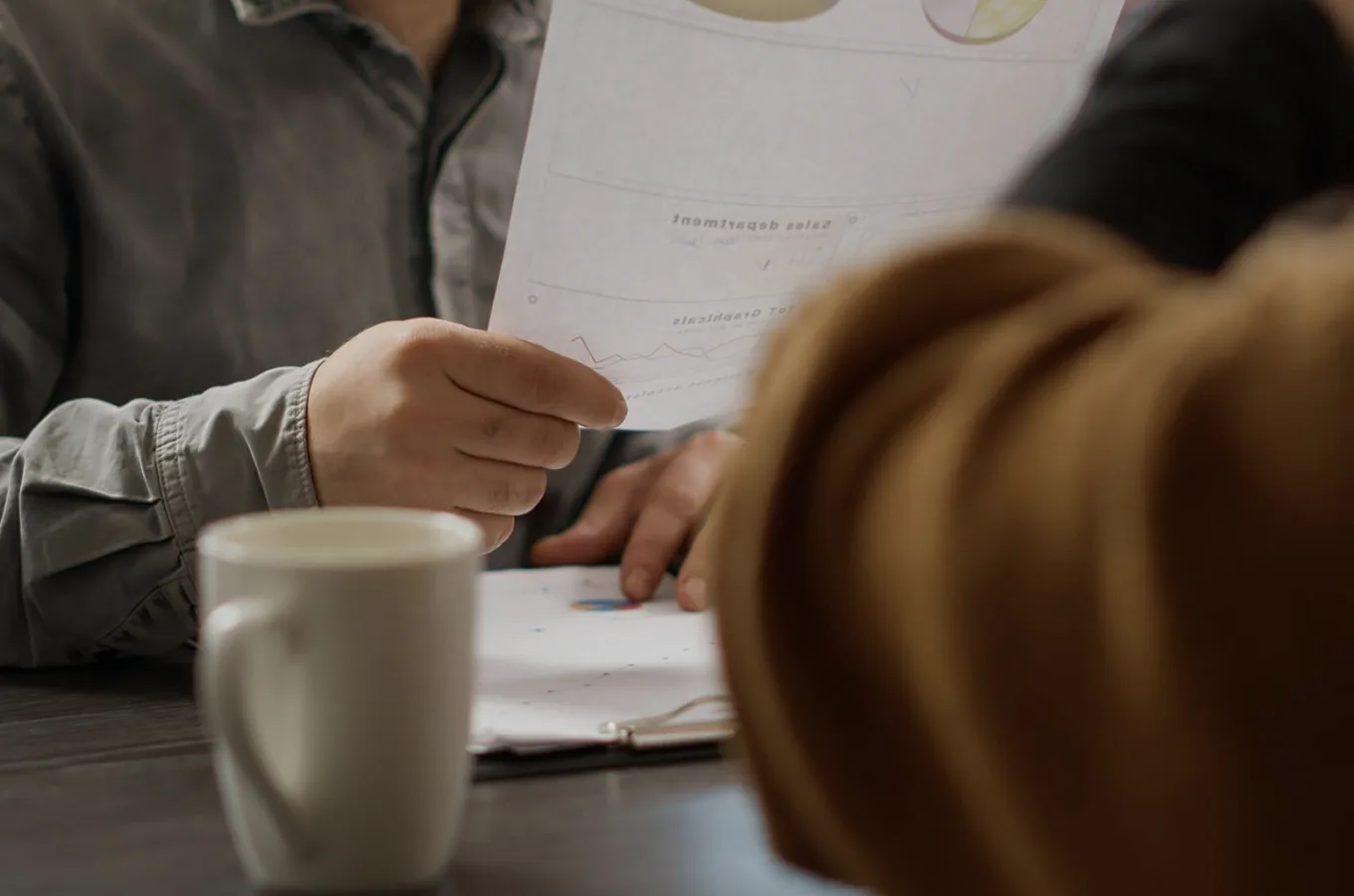 Two people at a table, one holding a printed chart across from the other, with a coffee mug in the foreground