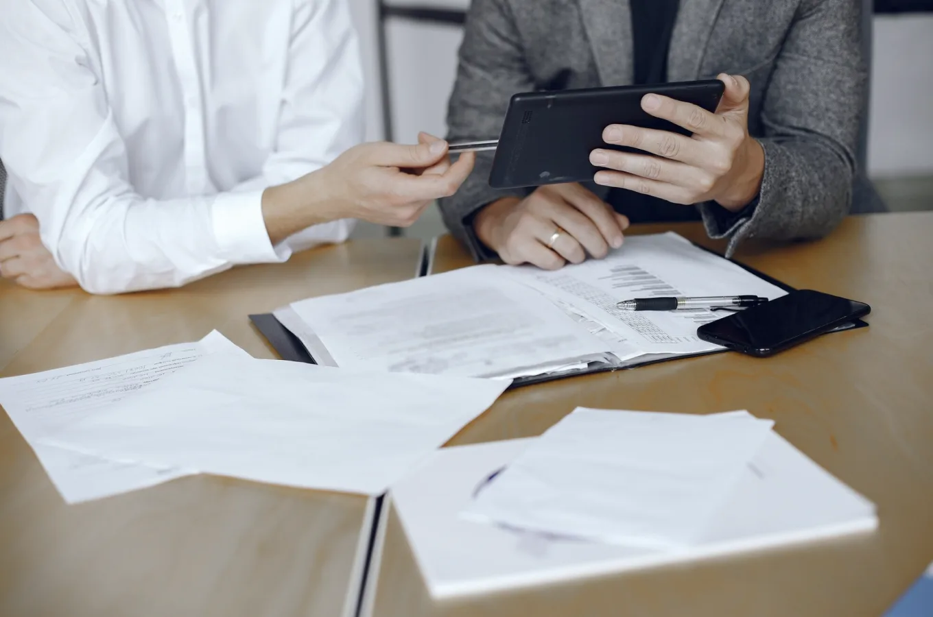 Two people seated at a table with documents, one holding a tablet while the other points at it with a pen