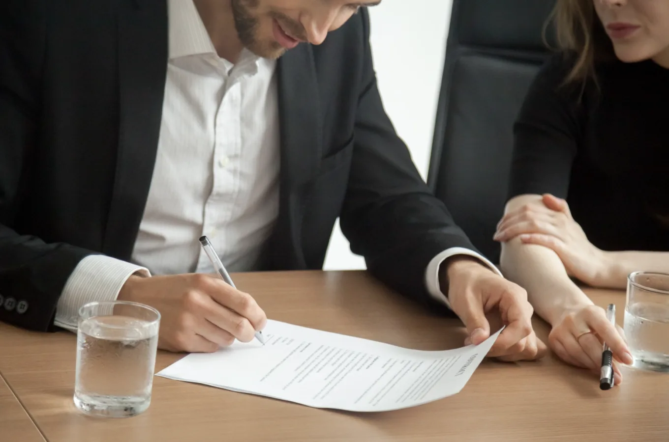 A man in a suit signing a document at a conference table, with a woman seated beside him and glasses of water on the table.