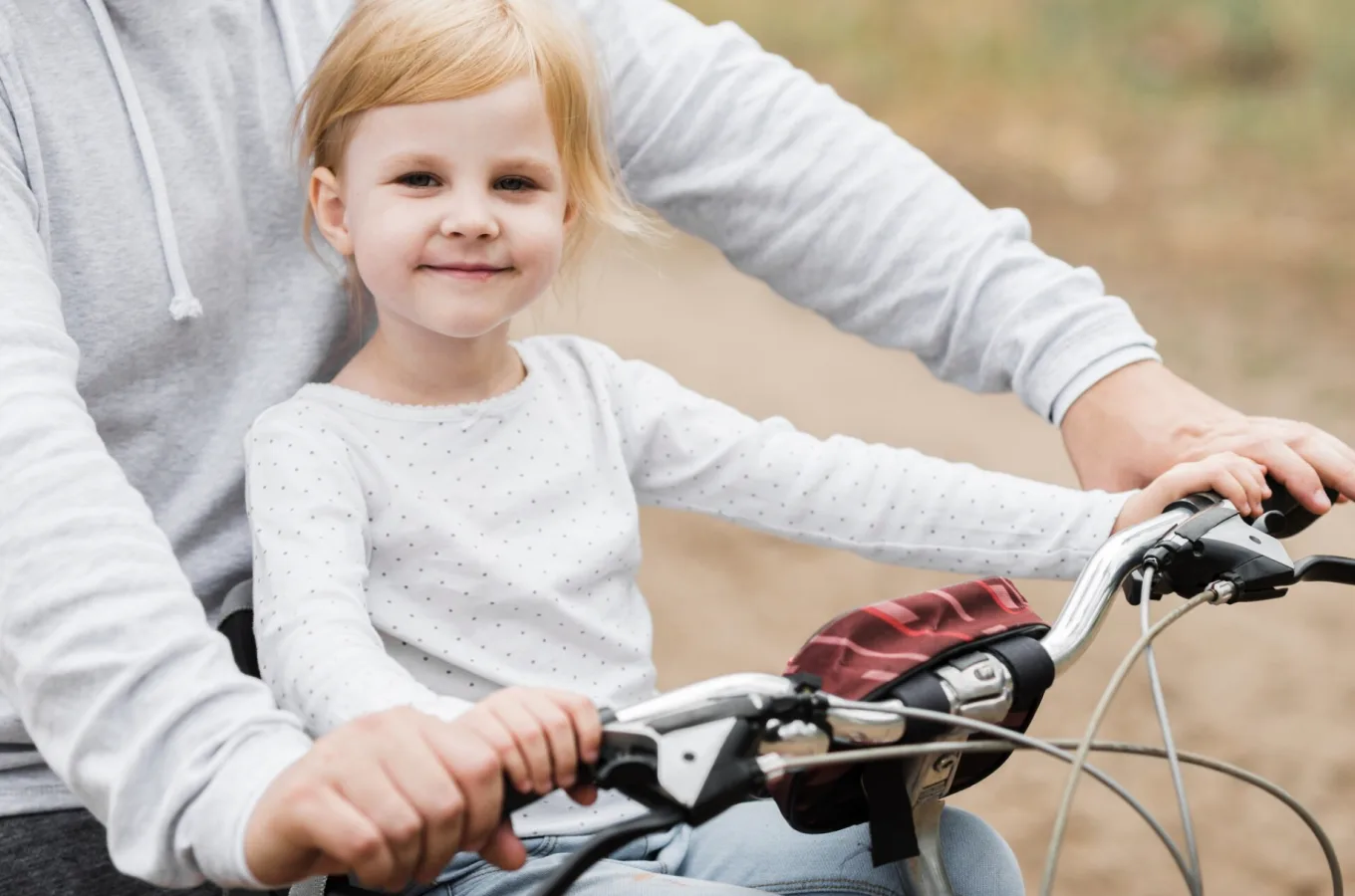 An adult in a grey sweatshirt steadying the handlebars of a bicycle while a smiling young child sits on it.